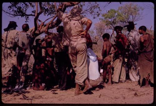 O.P.M. Prozesky, Wulf Haacke, and other expedition members standing with a group of people watching the cutting up of an ostrich, seen from behind