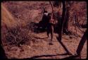 Little girl walking with an ostrich feather in her hair, women and children sitting in a patch of shade behind her