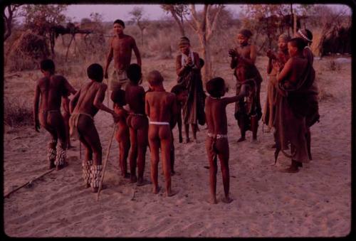 Adults gathering for a curing dance, women clapping and children dancing and two boys wearing rattles on their legs