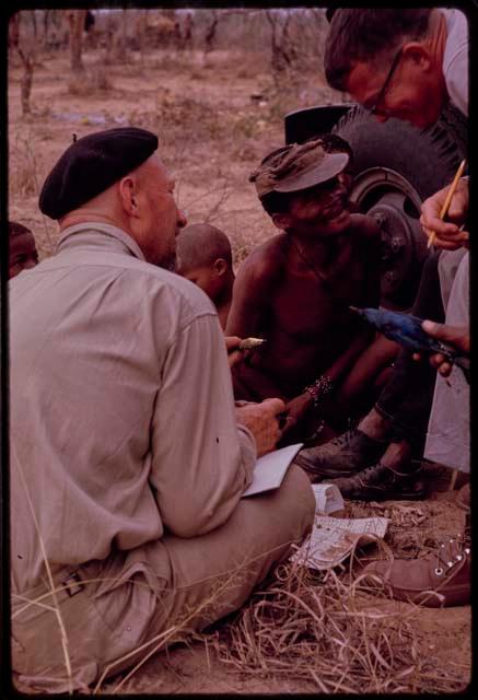 Group of people sitting with O.P.M. Prozesky, Nicholas England, Dabe, and Lorna Marshall and identifying birds; a man holding a bird and Nicholas England listening to his pronounciation, close-up