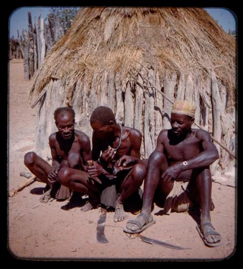 Three men with snuff sitting in front of a hut