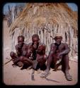 Three men sitting in front of a hut and taking snuff