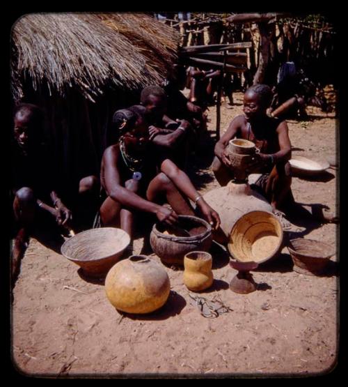 Group of women sitting in front of a hut with bowls in front of them