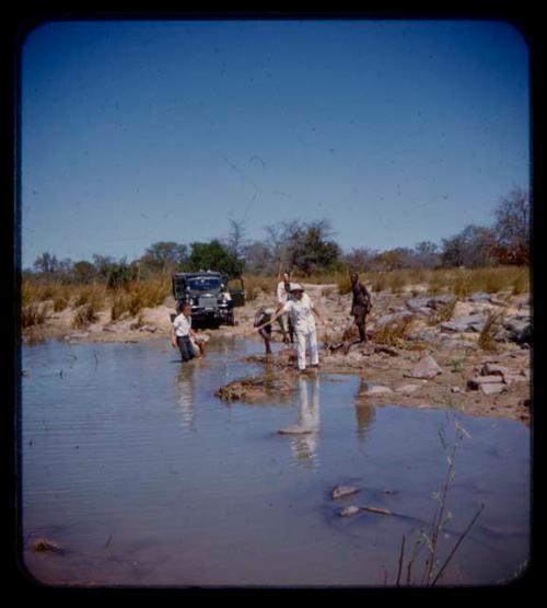 Expedition members standing in a river and on the bank, expedition truck on road