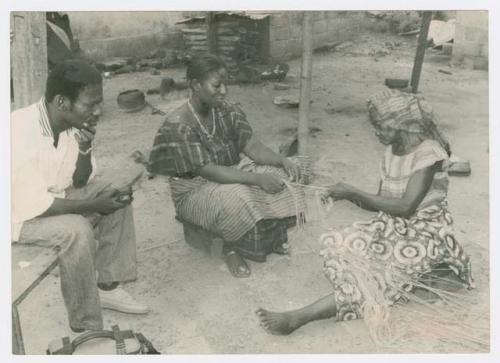 Two women sitting on the ground and weaving. Man sitting on bench.