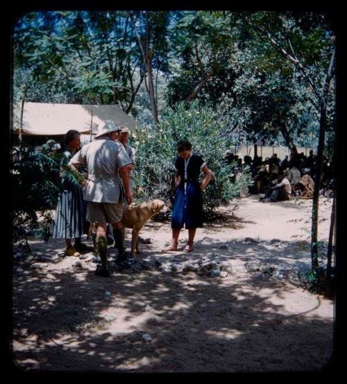 Lorna Marshall, John Marshall, and Elizabeth Marshall Thomas standing with a man and dog, a group of people sitting in the background