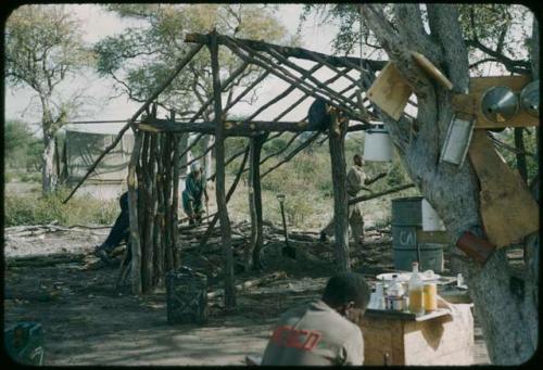 Expedition member sitting in front of a cookhouse, with /Gunda and two other men working in the background