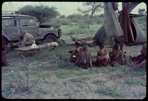 Group of people sitting, with Laurence Marshall sitting next to the expedition Land Rover near them, tent in the background