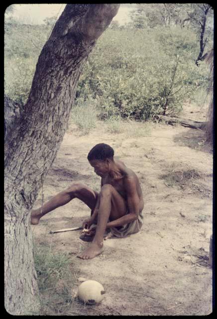 /Ti!kay working under a tree near his skerm, with an ostrich eggshell on the ground next to him, close-up