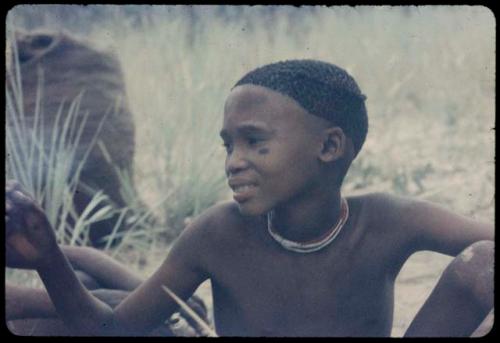 Child sitting, with a large bag, probably filled with mangetti nuts, in the background