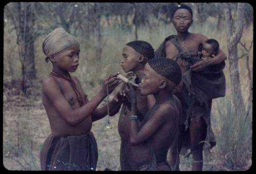 N!ai giving a pipe to a boy, with another boy standing next to them, !Ungka carrying a baby behind them