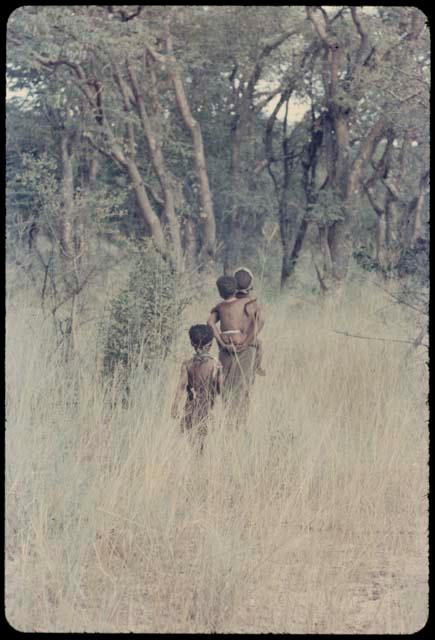 Woman walking, carrying Di//khao on her back, with !Ungka Norna following, view from behind