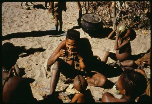 Woman sitting with a group of people, with a child drinking juice from a tsama melon behind her (copy of slide 11E-1)