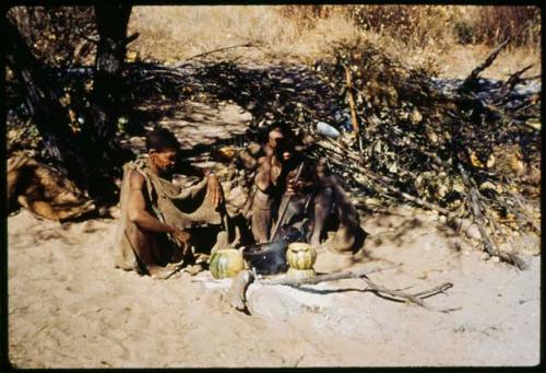 Two women sitting next to a fire, cooking, with tsama melons on the ground next to them, used as double-boilers (copy of slide 11E-241)