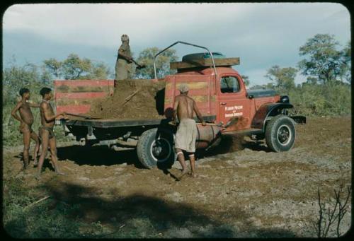 /Ti!kay and another man shoveling fertilizer from the back of the expedition truck, with two men standing next to the truck