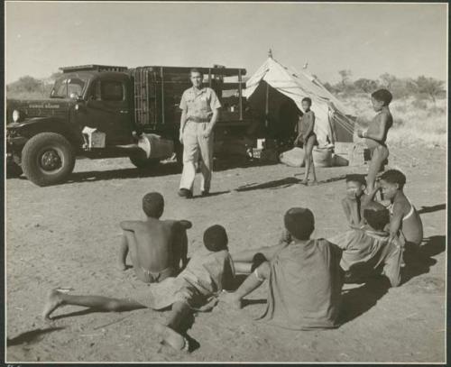 Group of people sitting and standing, with Heiner Kretzchmar walking in front of an expedition truck in the background (print is a cropped image)