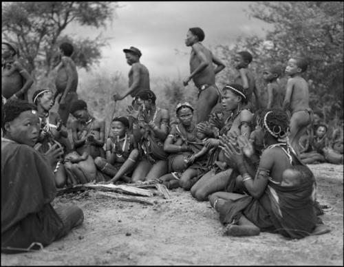 Group of women sitting in a circle singing and clapping, with men dancing behind them in a curing dance