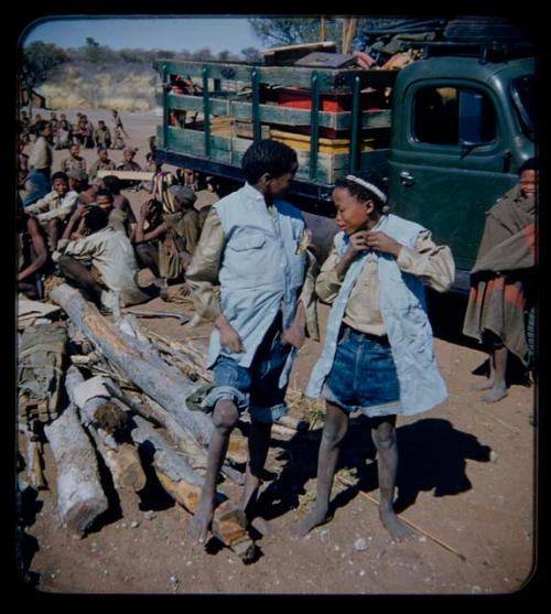 Expedition: Boy and girl wearing Western clothing, with a woman wrapped in a blanket standing by an expedition truck behind them, group of people sitting in the background