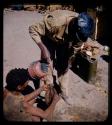 Expedition: Expedition member bending over to pour water from a kettle into an animal skin bag held by a seated man