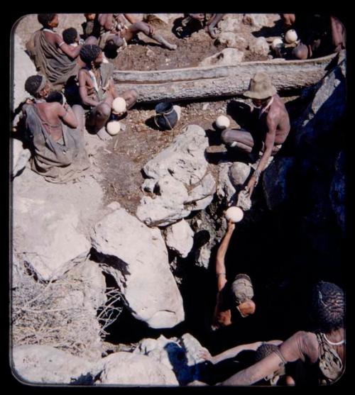 Waterhole: Person handing an ostrich eggshell filled with water to another, with other people in the background