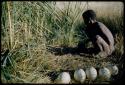 ≠Gao (leader of Band 10) filling ostrich egg shells with water at a waterhole, with five ostrich egg shells with stoppers and an Ovambo pot on the ground next to him