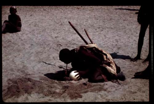 Person drinking water from a hole in the sand, holding an ostrich eggshell