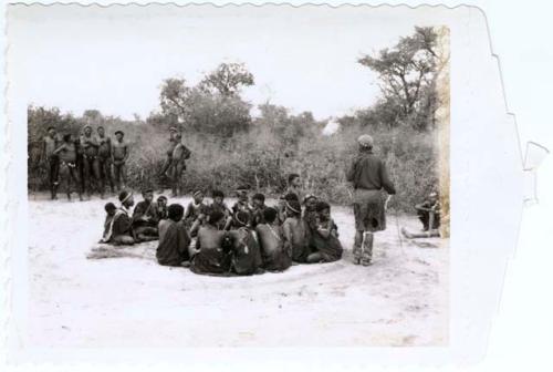 Man wearing Western clothes and dance rattles, dancing around a group of women and children sitting, with a group of men standing in the background