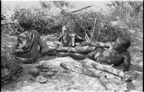 !Gai lying beside his fire, Tsekue and N!whakwe sitting beside him