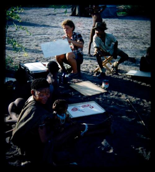 Expedition: Elizabeth Marshall Thomas holding a drawing and Ngani looking at it over his shoulder, with a woman and children with drawings and painting tools in the foreground