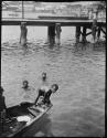 Boys diving for pennies next to a boat, with a bridge in the background