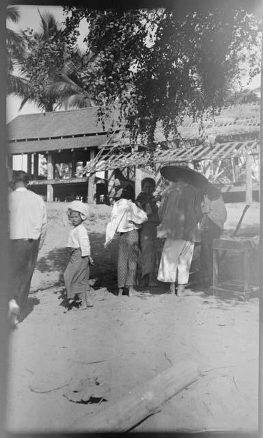 People standing under a tree, with a large building on stilts in the background