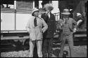 Three men dressed in Western clothing, smoking cigars, standing in front of a train