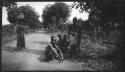 People sitting and standing at the edge of a road, with a woman carrying a bundle on her head walking near them