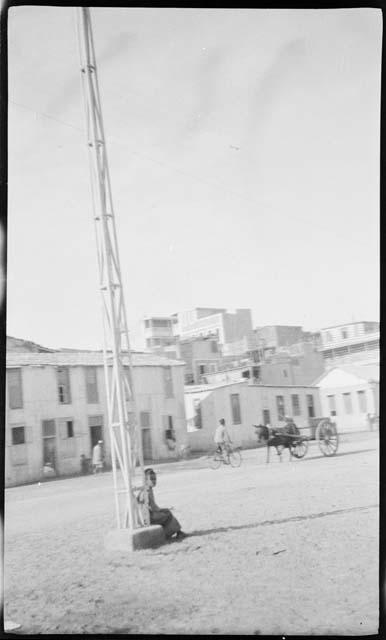 Person sitting against a tower, with bicycle, ox cart and buildings in the background