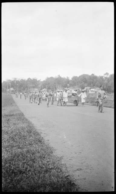 People pulling carts down a road, with men in uniforms walking next to them