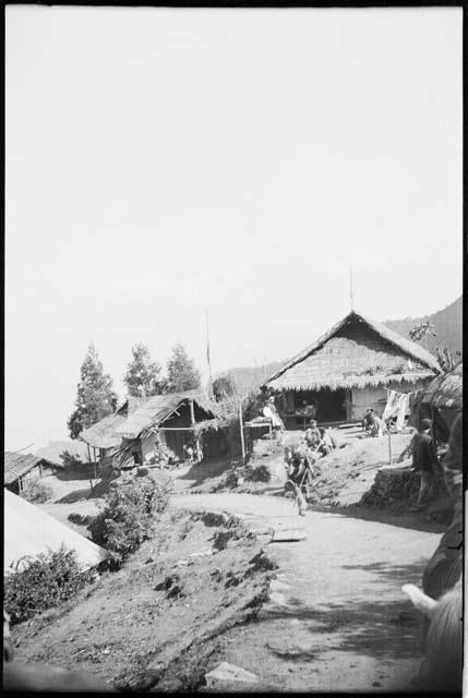 Man carrying bundles down a road, with people sitting in front of thatched-roof buildings in the background