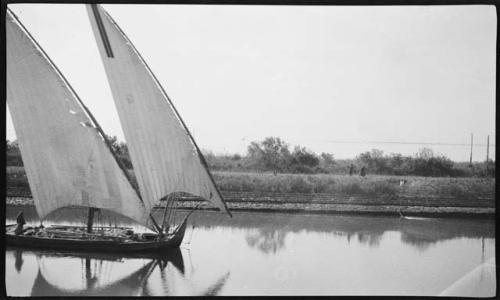 Person standing on a felucca (boat), with people walking along shore in the background