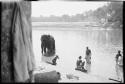 People sitting at the edge of the water, and standing in the water next to an elephant