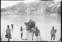 People standing in the water next to an elephant, with a ghat in the background