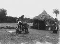 Men cooking in iron pots over a fire in the expedition camp, with a hut in the background