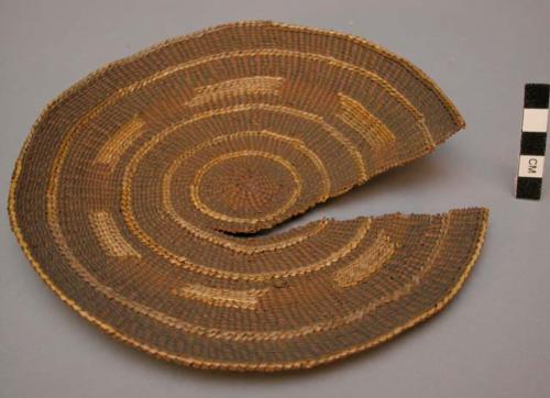 Circular basketry mat of yellow and brown grasses, torn