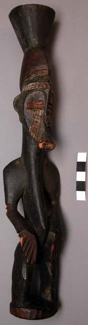 Elongated carved figure, wood, with hands resting on knees, scarified face and p