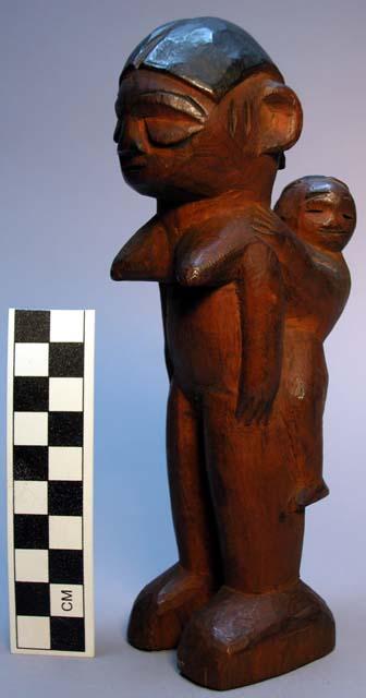 Wooden figure, mother carrying child lengthwise on her back.  Contrasting brown